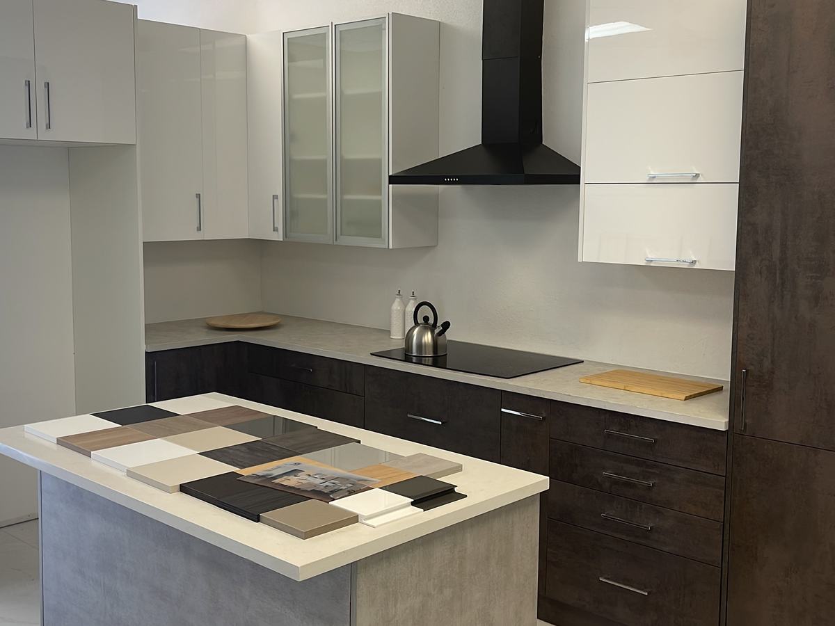 Cocina de exhibición con gabinetes altos blancos brillantes, puertas de vidrio esmerilado, campana negra, gabinetes bajos con veta de madera oscura y muestras de acabados sobre la isla