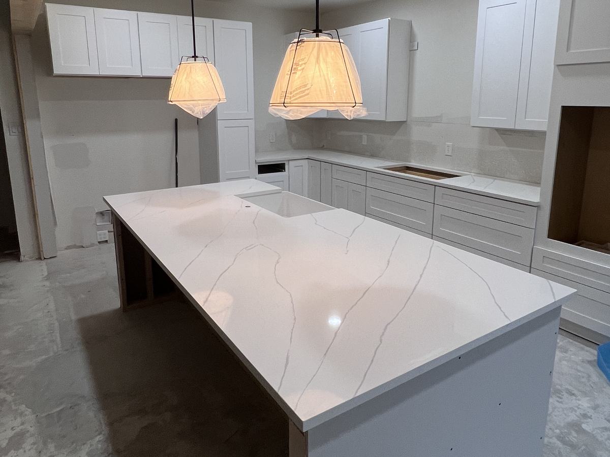 Isla en cascada de cuarzo blanco con vetas grises bajo dos lámparas colgantes, en una cocina con gabinetes blancos estilo Shaker, antes de instalar el piso