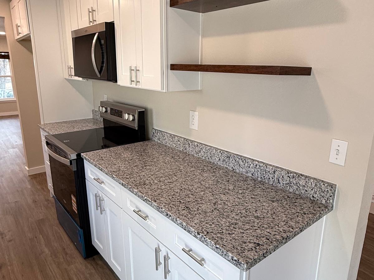 White Shaker-style cabinets with a speckled gray granite countertop, a stainless range and microwave, and two dark wood floating shelves